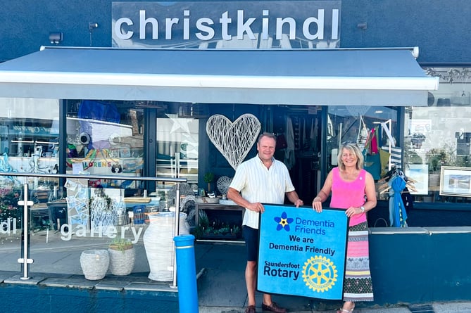 John Walker-Smith, the Treasurer of Tenby Memory Café and lead volunteer in the Dementia Friendly Saundersfoot project, is seen with Lesley displaying the new blue floor mat, kindly supplied by Saundersfoot Rotary Club, which is now in place welcoming customers at Christkindl.