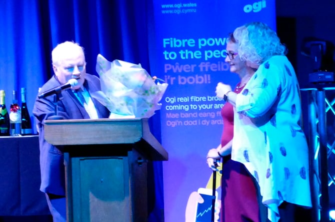 Tenby Observer Kindness in the Community event organiser Carolyn Cox is accompanied onto the stage by Heather Wood, Sales Manager, The Forester, Forest Review & The Ross Gazette, to receive a ‘thank you’ bouquet from Mick O’Reilly, Regional Editor, Cambrian News and Tenby Observer.